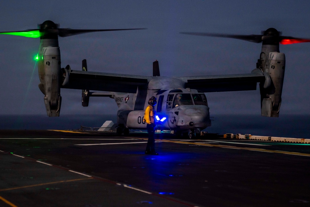 A tiltrotor aircraft sits on the flight deck of a ship, its propellers illuminated by red and green lights, as a sailor wearing a yellow jacket and a helmet looks on in dark conditions.