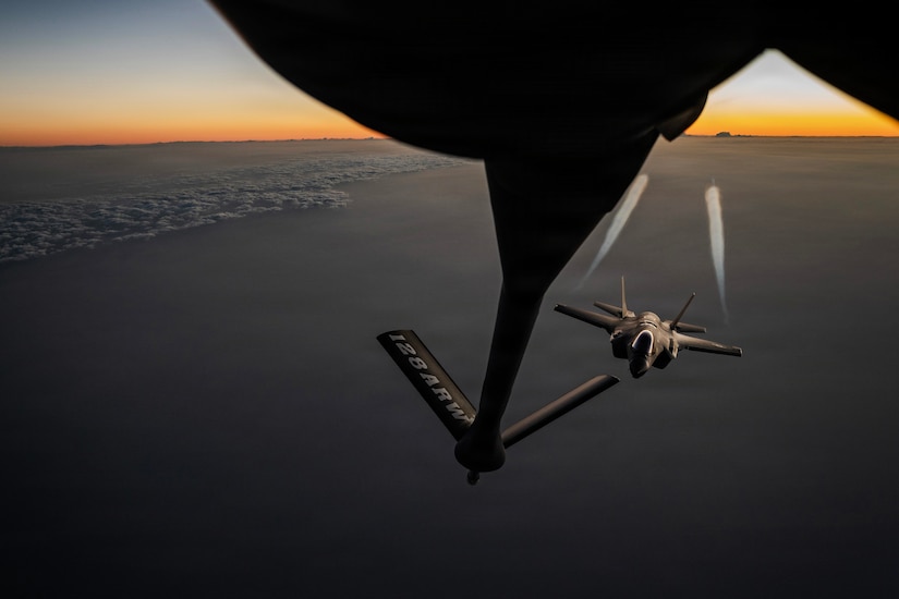 A flying jet with contrails from each wing approaches a partially visible aircraft with a boom extended from the rear, with clouds below and an orange and light blue sky in the background.