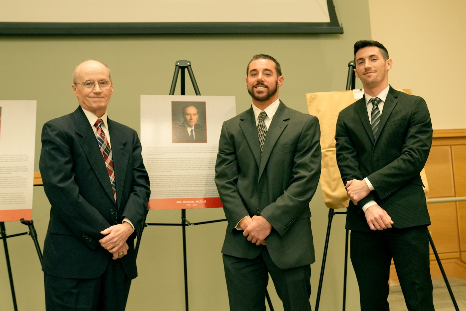 three men in suits stand on stage next to plaque