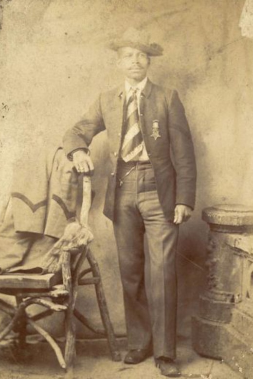 A black and white photo of a man in a suit posing for a photo while leaning on a chair. The Medal of Honor is pinned on the man's jacket.
