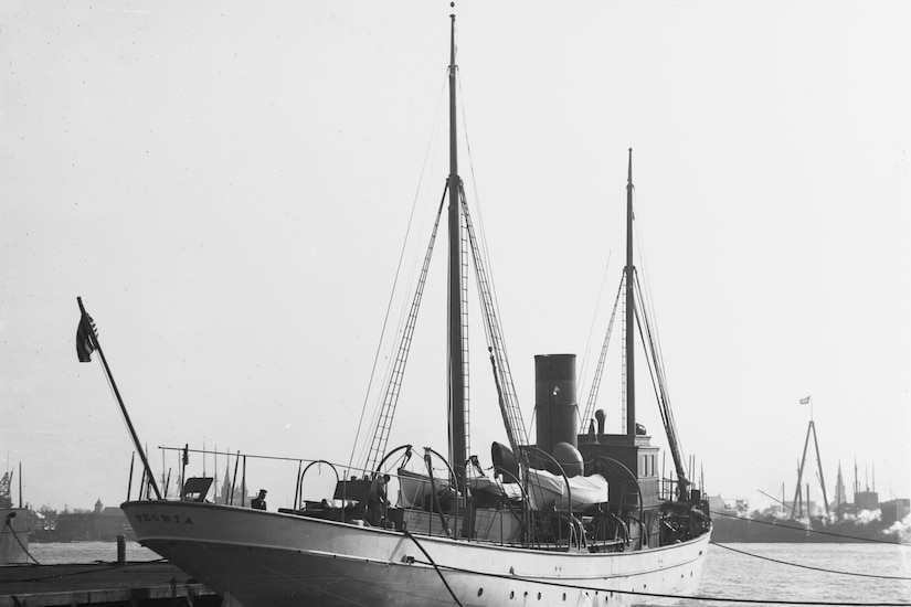 A black and white photo of a small military ship tied to a dock in a harbor.