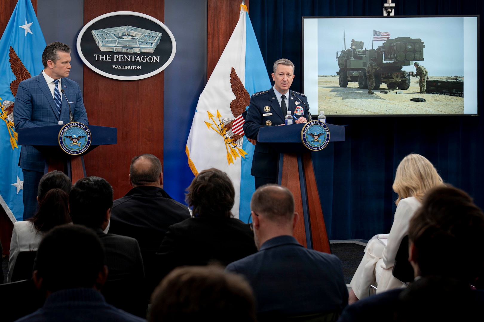 A man in business attire and a man in a formal military uniform stand at lecterns and speak to an audience. Behind them is a sign reading, "The Pentagon, Washington," and various flags. To the right, an image of a military vehicle is displayed on a monitor.