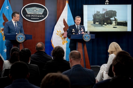 A man in business attire and a man in a formal military uniform stand at lecterns and speak to an audience. Behind them is a sign reading, "The Pentagon, Washington," and various flags. To the right, an image of a military vehicle is displayed on a monitor.