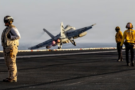 An aircraft lifts off the deck of a carrier. Two individuals in yellow jackets stand on the deck. Another individual in a beige vest wears a helmet that reads QAS.