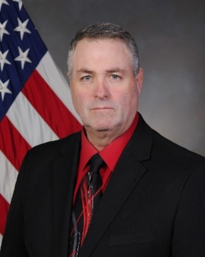 Man wearing a red shirt and black suit stands near an American flag