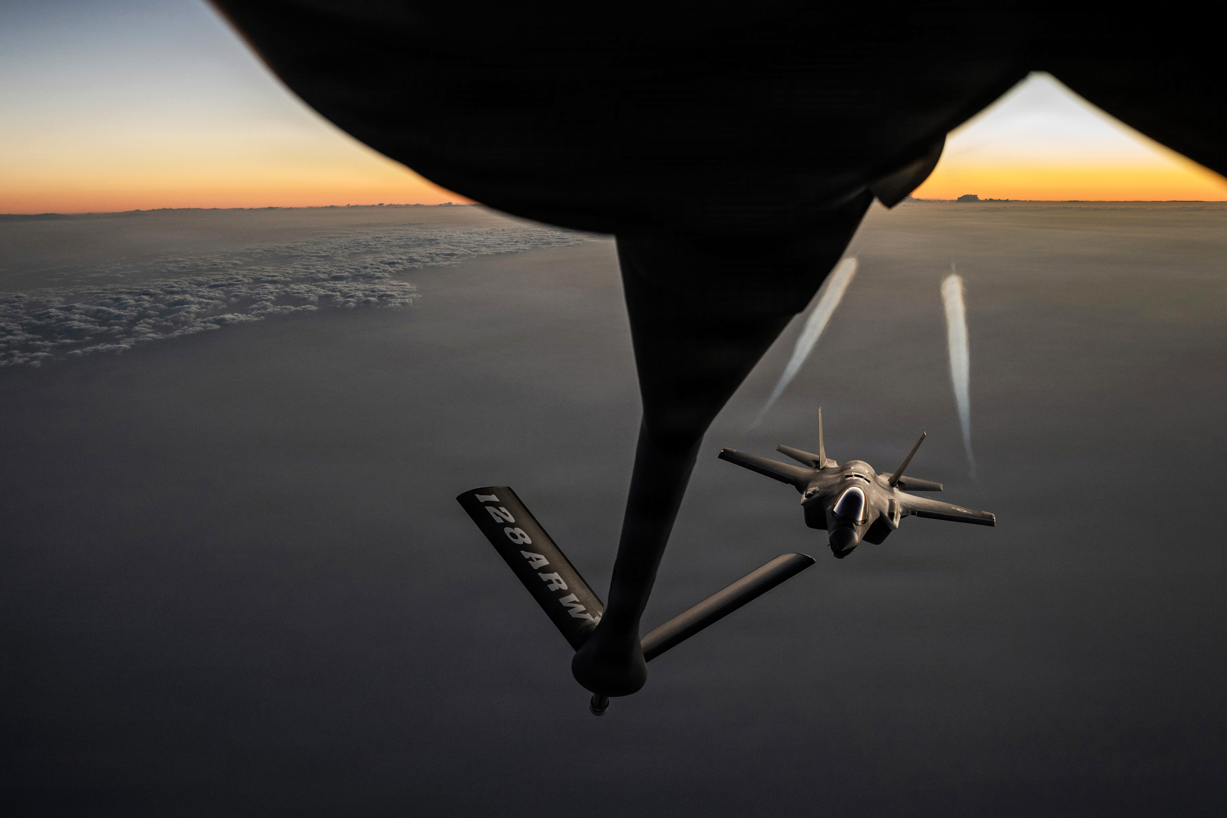 A jet's wings create contrails as the aircraft flies below and behind a fuel boom from another aircraft.