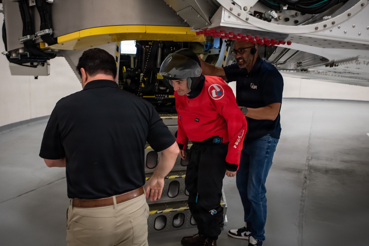 A man wearing a red jacket, black pants and a helmet exits a training simulation device, assisted by two men on either side of him.