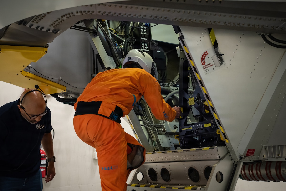A man wearing an orange flight suit climbs into a centrifuge while another man in civilian attire stands to the left of him.
