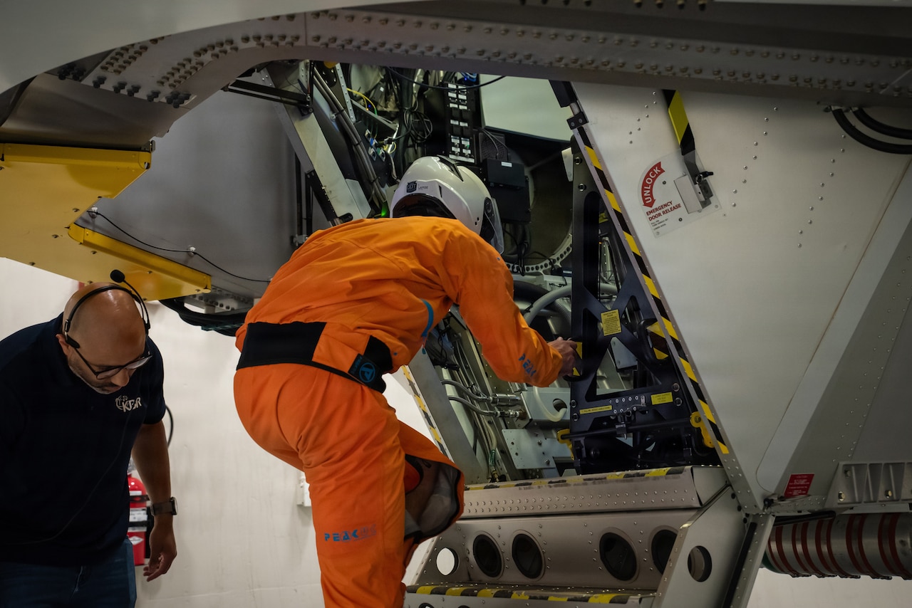 A man wearing an orange flight suit climbs into a centrifuge while another man in civilian attire stands to the left of him.