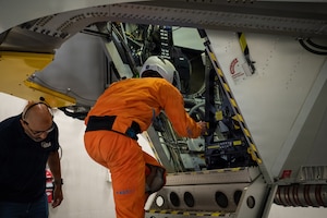 A man wearing an orange flight suit climbs into a centrifuge while another man in civilian attire stands to the left of him.