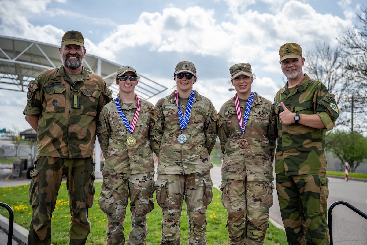Three women and two men pose for a group photo. The three women, wearing military camouflage uniforms, have gold, silver and bronze medals around their necks