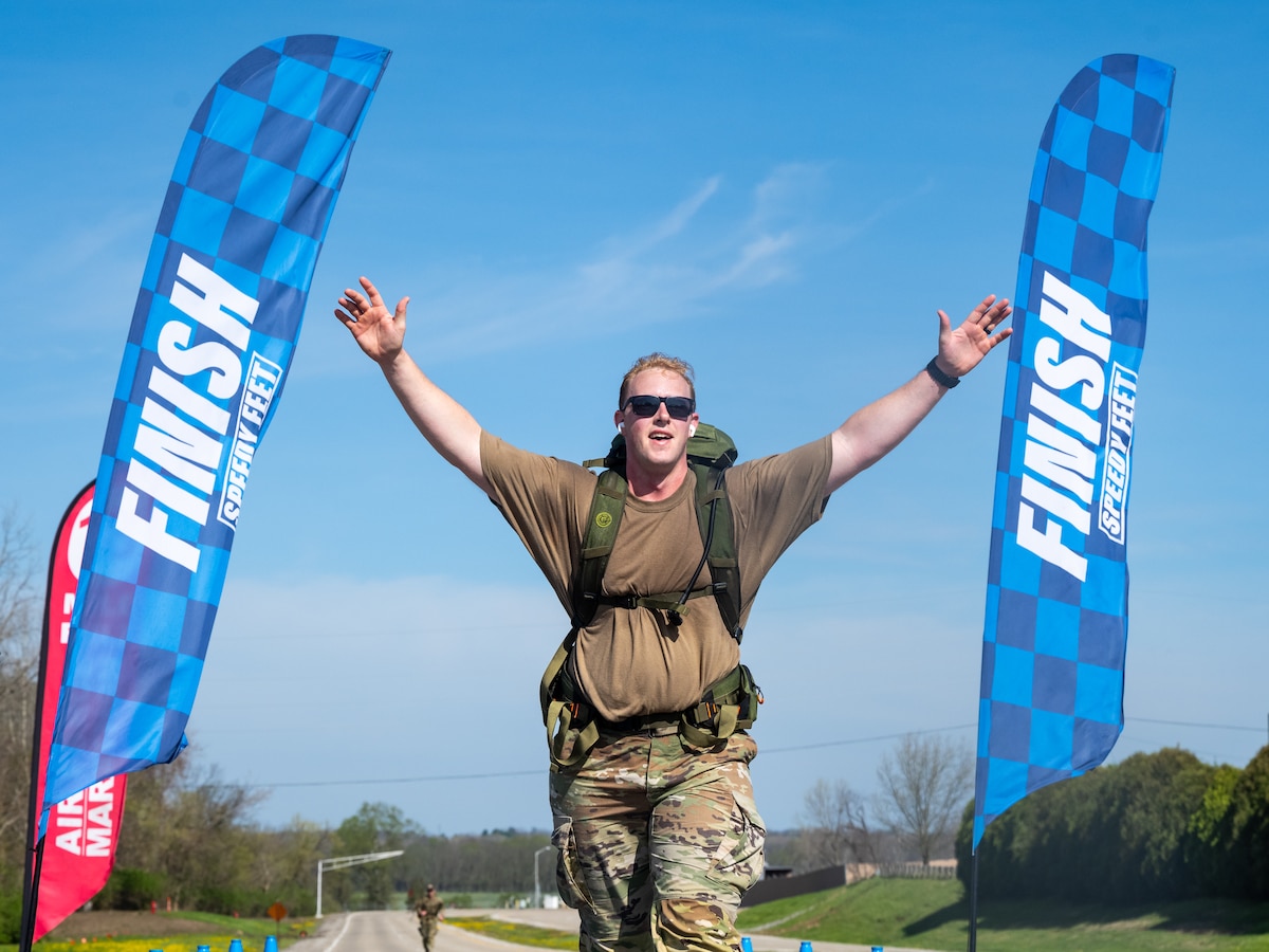 A man, wearing brown t-shirt, military camouflage pants, black sunglasses and backpack, raises is arms in victory. Two vertical blue banners, reading FINISH, are on either side