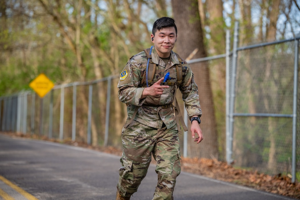 A young man, wearing military camouflage uniform and backpack, gives a peace sign while smiling at the camera and running down a paved road alongside a silver chain link fence