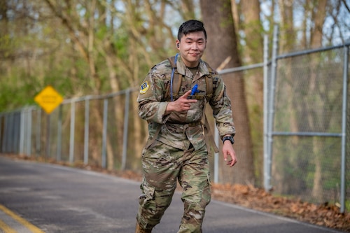 A young man, wearing military camouflage uniform and backpack, gives a peace sign while smiling at the camera and running down a paved road alongside a silver chain link fence