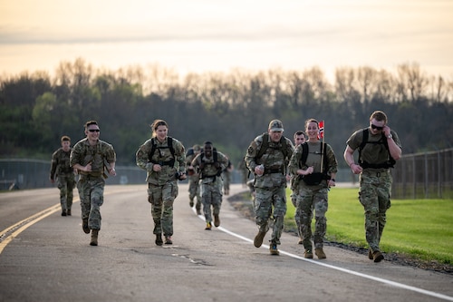 A group of men and women, wearing military camouflage uniforms and wearing large backpacks, run down a paved road