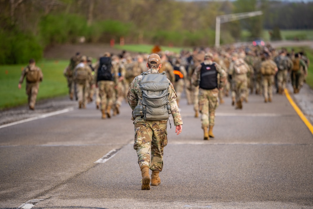 A behind view of a large group of people, most wearing military camouflage uniforms and large backpacks, marching down a paved road