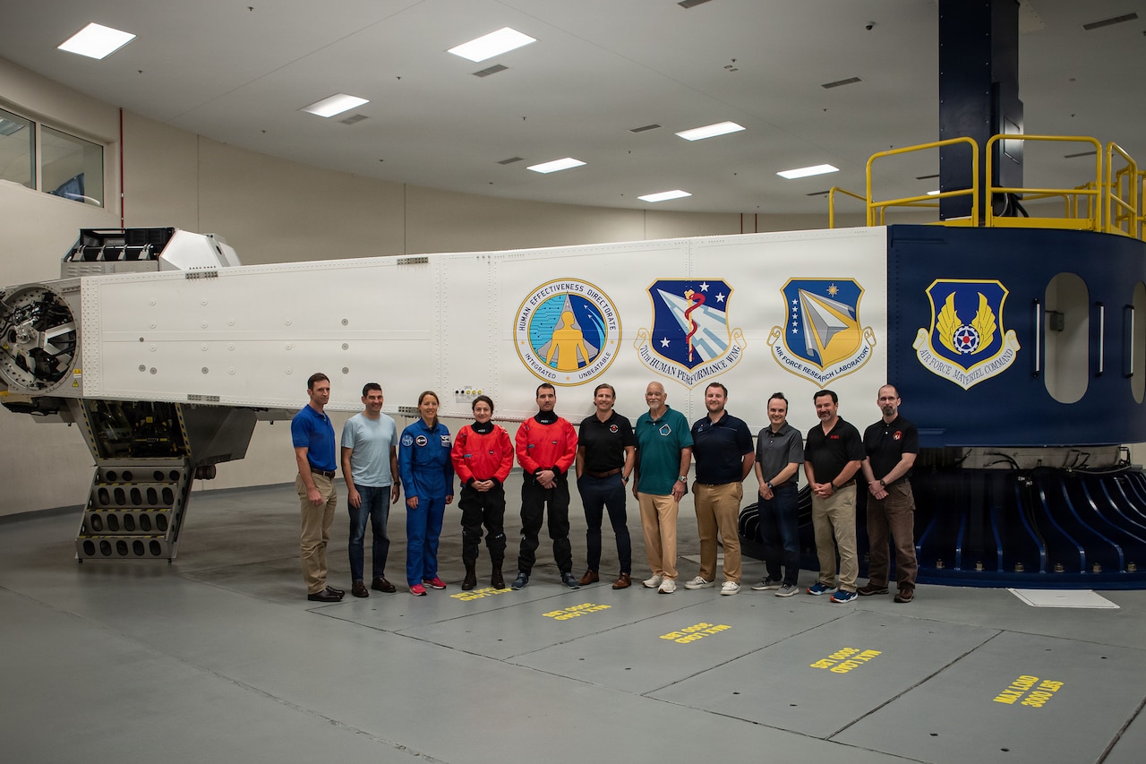 A group of people wearing civilian attire, and one wearing a blue flight suit, pose for a photo in front of a centrifuge.