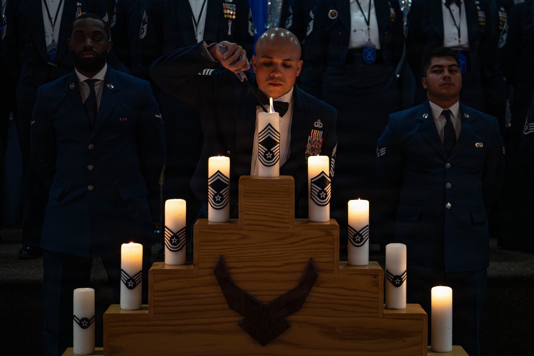 An airman lights candles on a candelabra as fellow airmen watch in formation in the dark background.
