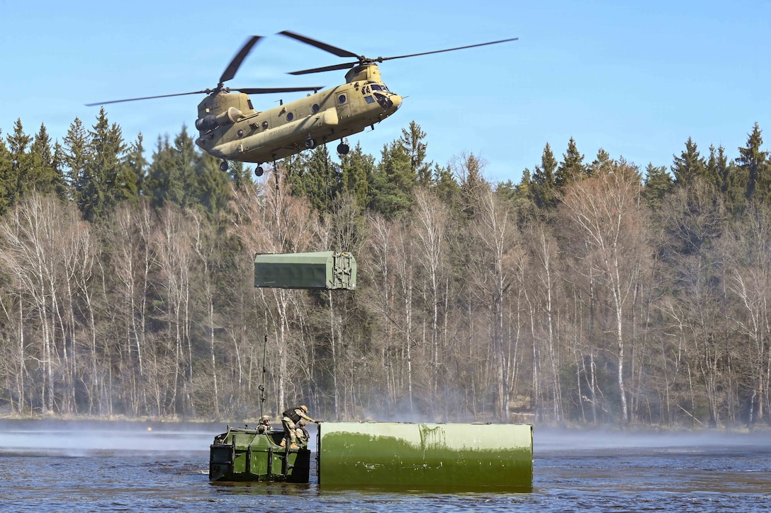 Soldiers climb a green makeshift bridge in a body of water as a helicopter hovers carrying a piece of equipment in a blue sky with woods in the background.