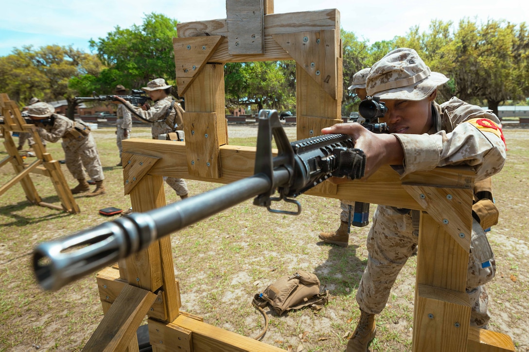 A Marine Corps recruit leans on a wooden structure while looking through a rifle's scope in a field during the day.