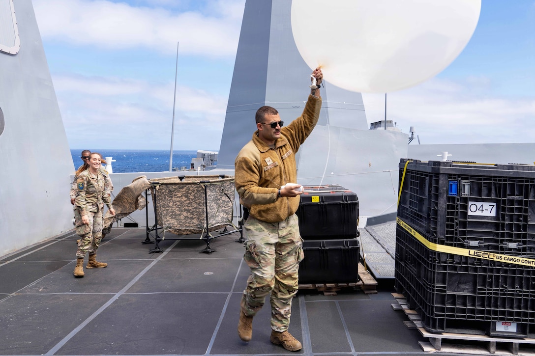An airman holds a large white balloon aboard a ship at sea as two service members walk nearby.