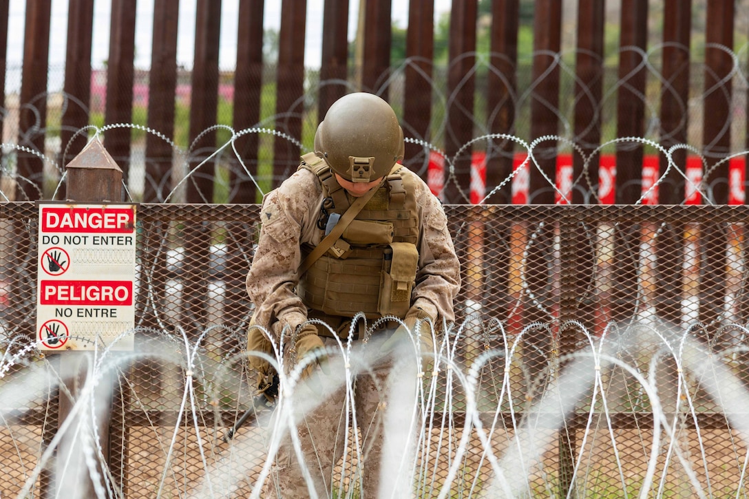 A Marine in tactical gear works on concertina wire in front of a metal fence.