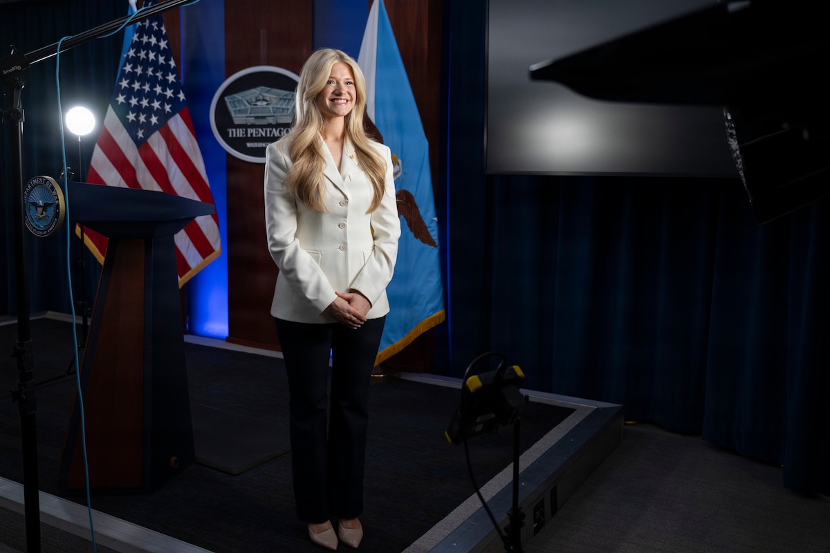 A woman in business attire stands and smiles with lights and camera equipment around her; behind her is an American flag and a sign on the wall that reads, "The Pentagon."