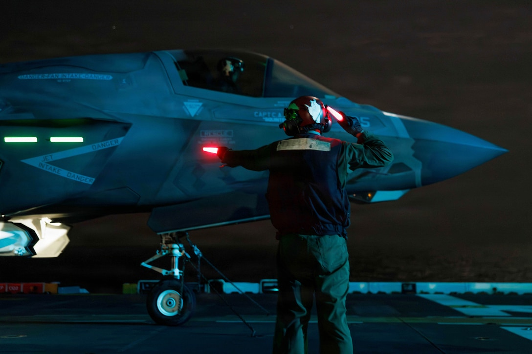 A sailor holds two red batons next to an aircraft aboard a ship in the dark.