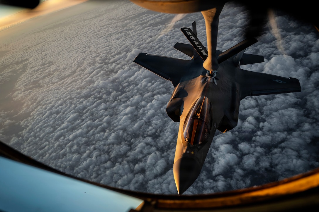 An aircraft extends its boom to another aircraft as they fly over clouds in a sunlit sky.
