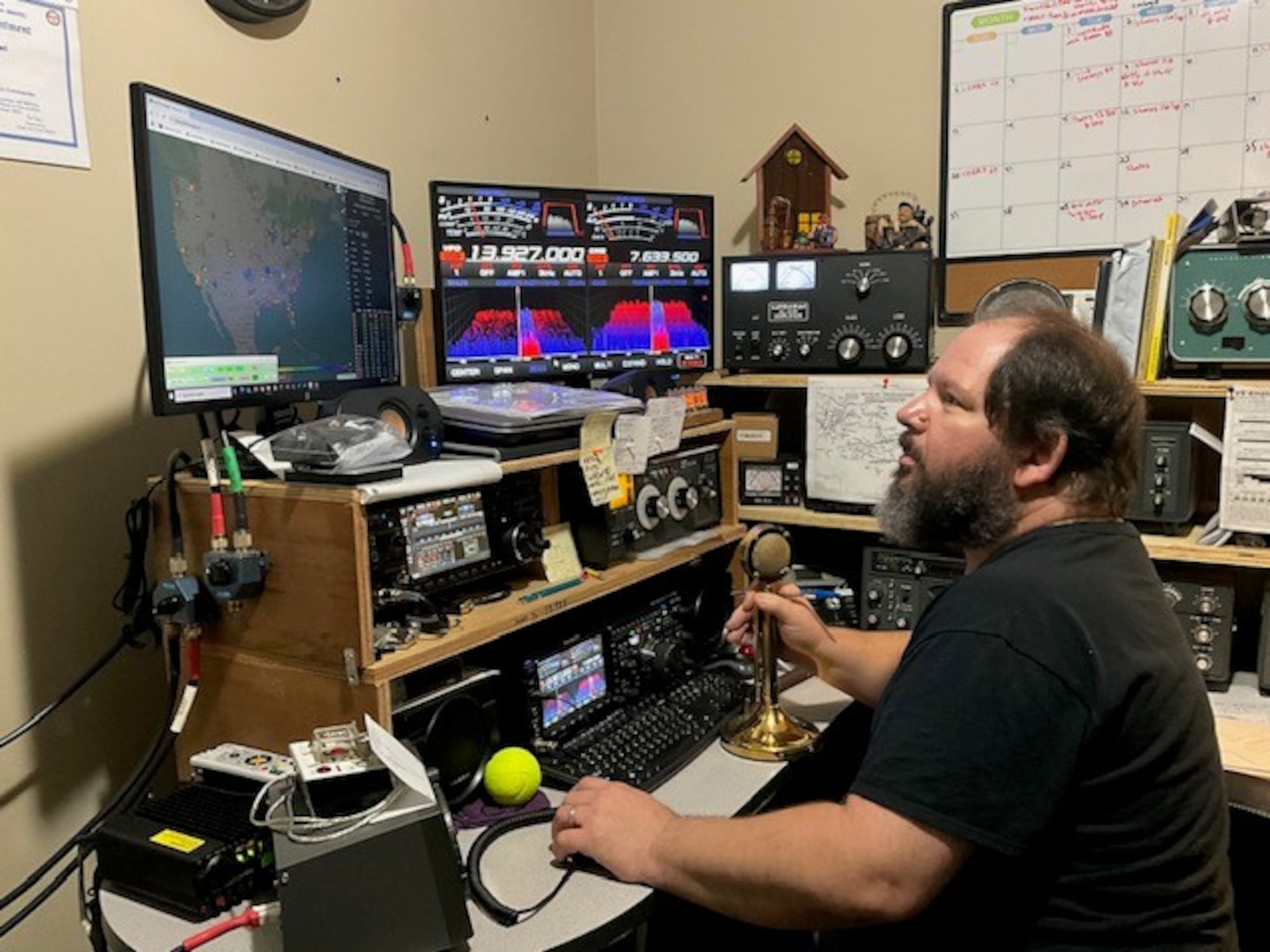 A man sits at a chair, staring up at a screen that shows a picture of the United States. To the right of that screen is an assortment of colors and lines. The man is also surrounded by radio equipment.