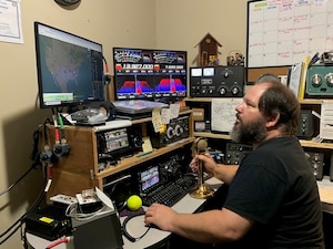 A man sits at a chair, staring up at a screen that shows a picture of the United States. To the right of that screen is an assortment of colors and lines. The man is also surrounded by radio equipment.