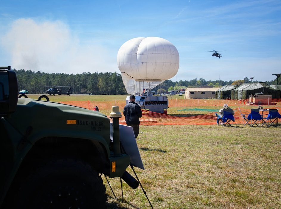 Military personnel and engineers collaborate in a field environment during the System of Systems Naval Integration Experiment (SoSNIE), operating communication equipment and digital systems under a camouflage net to test interoperability and command-and-control capabilities.