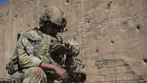 A man wearing a camouflage military uniform and helmet sits in a desert environment and looks at a handheld device; a person in similar attire is behind him doing the same.