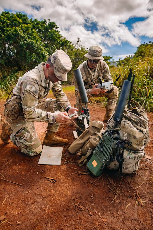 Two people wearing camouflage military uniforms crouch in the dirt to assemble military equipment under a blue sky with clouds, and trees in the background.
