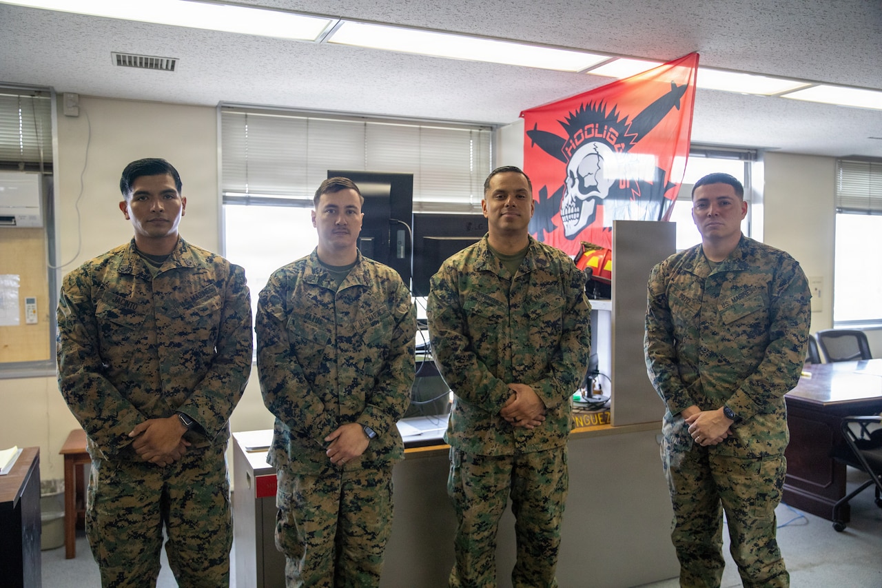 Four Marines in camouflage uniforms stand and pose for a photo in an office-like setting, with a red flag with a skull on it hanging in the background.