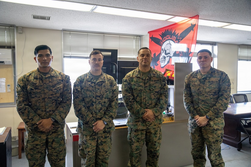 Four Marines in camouflage uniforms stand and pose for a photo in an office-like setting, with a red flag with a skull on it hanging in the background.