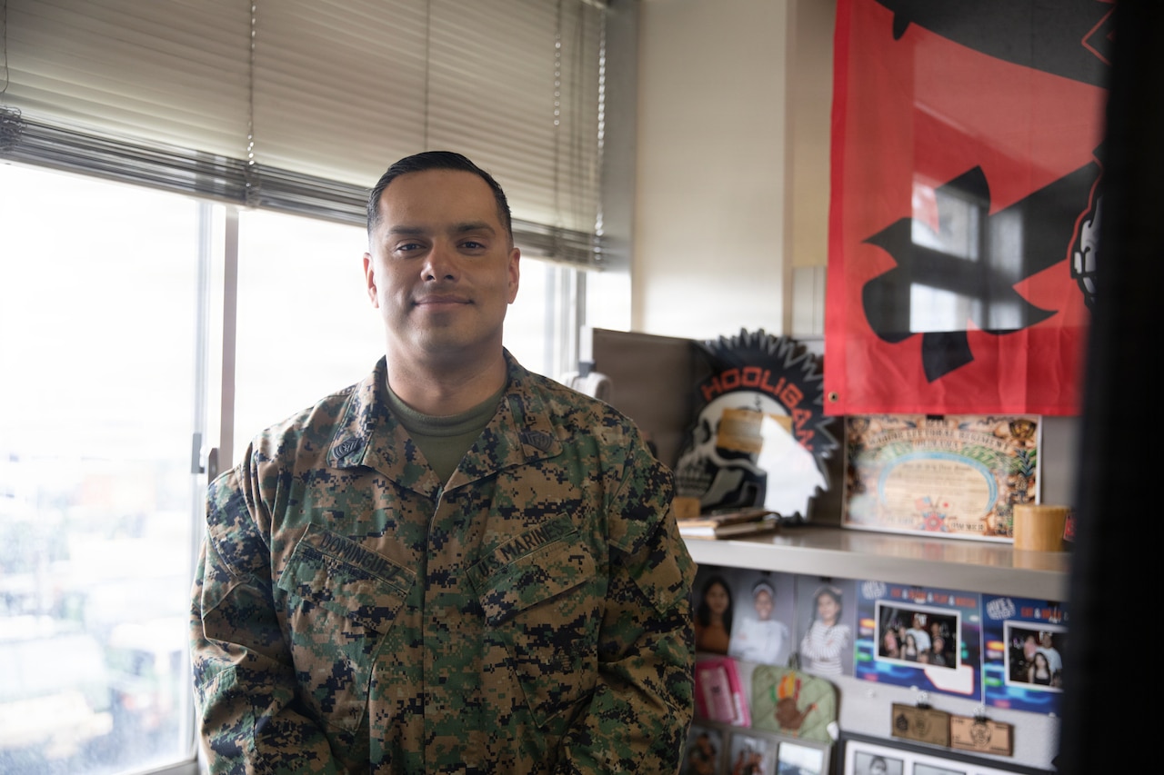 A Marine stands and smiles in an office-like setting, with a large window in the background.