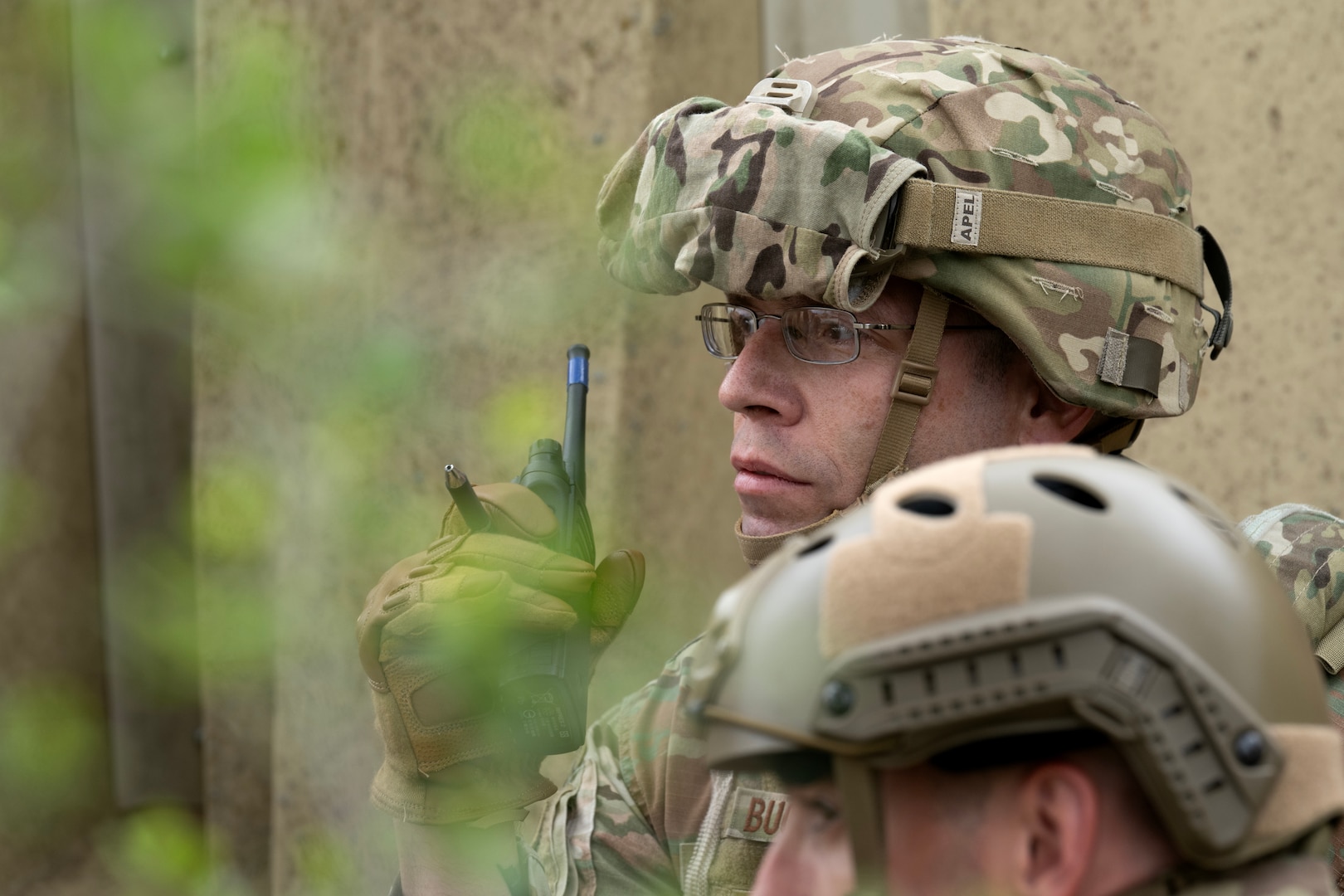 This is a close-up shot of a servicemember through the trees holding his walkie-talkie.