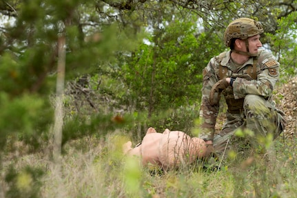Servicemember in a wooded area with trees is kneeling down next to a dummy.