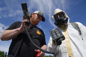 A man in civilian clothes and a person in a white hazmat suit controls a piece of equipment.