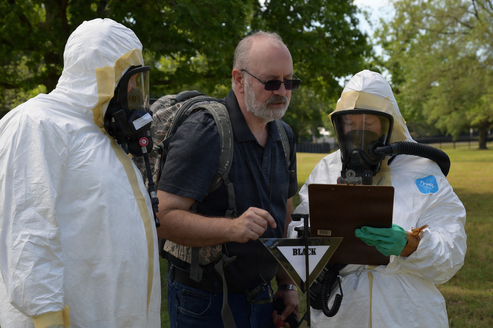 Two people in white hazmat suits review a chart with a man in civilian clothes in an open field.