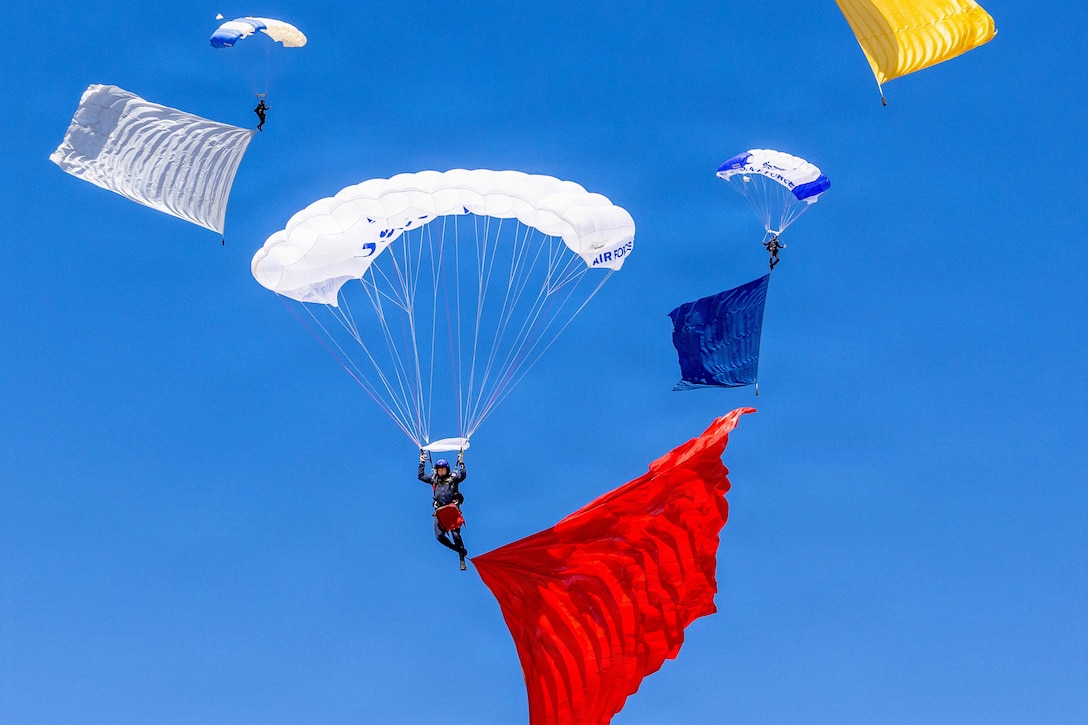 A few airmen attached to parachutes are seen in the air with a large piece of material in either red, white, blue or yellow attached to their feet and a clear blue sky in the background.