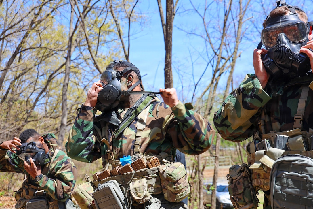 Three soldiers in camouflage uniforms secure gas masks outside, with trees and a blue sky in the background.