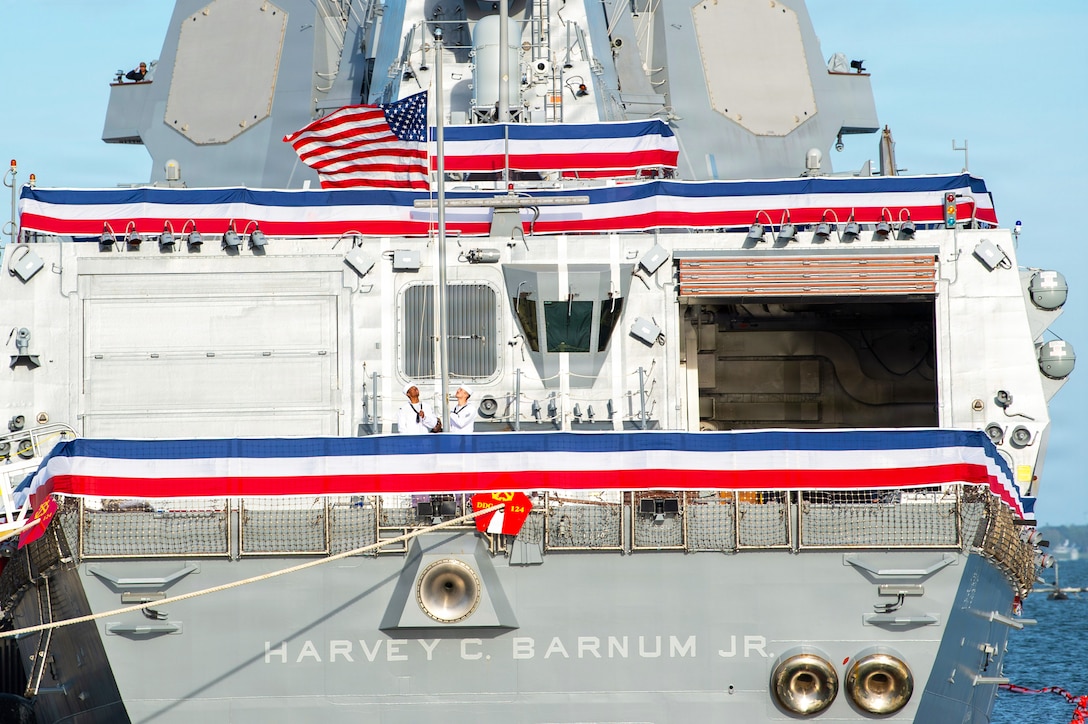 Two sailors at one end of a light gray military ship hold the ropes attached to an American flag on a flagpole, and three red, white and blue banners hang against the railings of different decks.