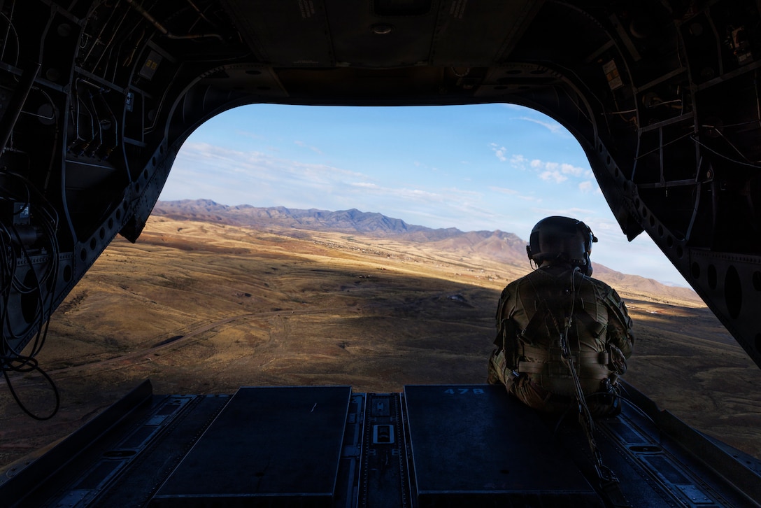 The back of a soldier is seen as he looks out at brown terrain and mountains below him while sitting in an open doorway of an aircraft.