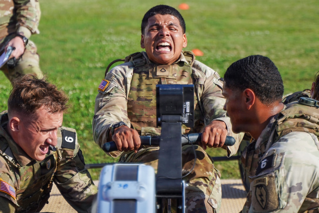 A soldier strains while using a rowing machine outside as two fellow soldiers kneel on opposite sides of the equipment.