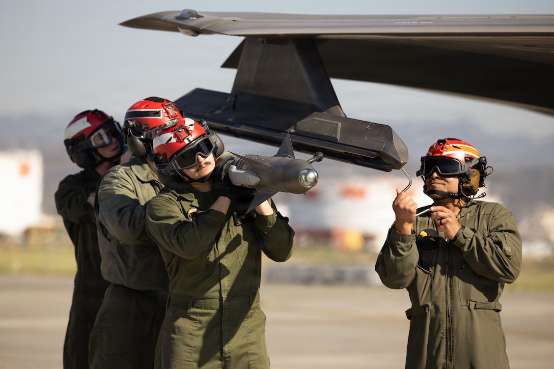 Three Marines in green jumpsuits and red helmets carry a missile as another Marine works on a partially visible aircraft during the day.