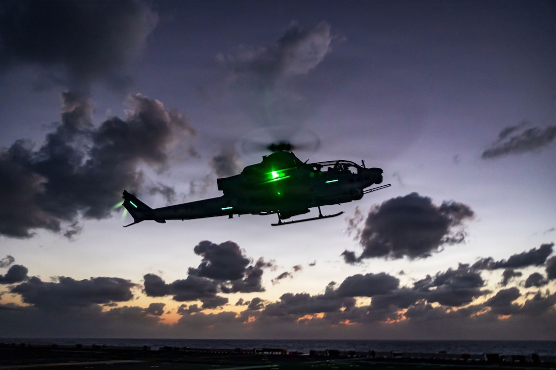 A helicopter illuminated by a green light flies in a dark purplish sky as the sunlight sits above the horizon.