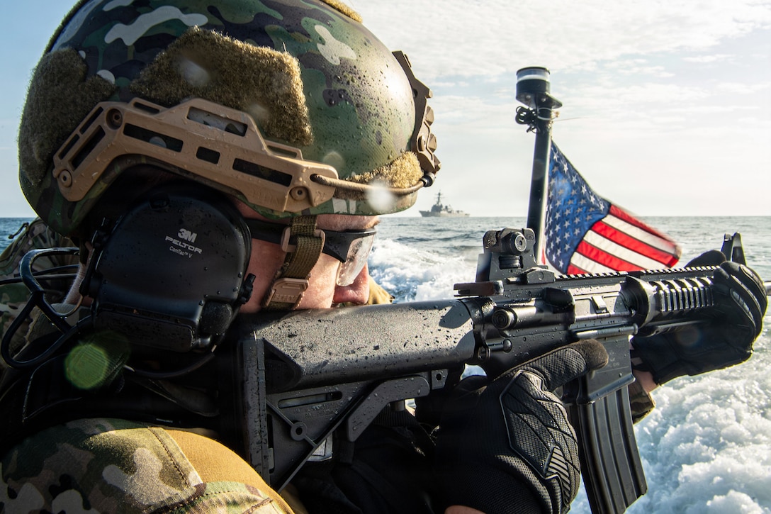 A close-up of a sailor in tactical gear aims a weapon near an American flag on a small boat at sea as a ship sails in the background during the day.