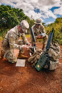 Two people wearing camouflage military uniforms crouch in the dirt to assemble military equipment under a blue sky with clouds, and trees in the background.